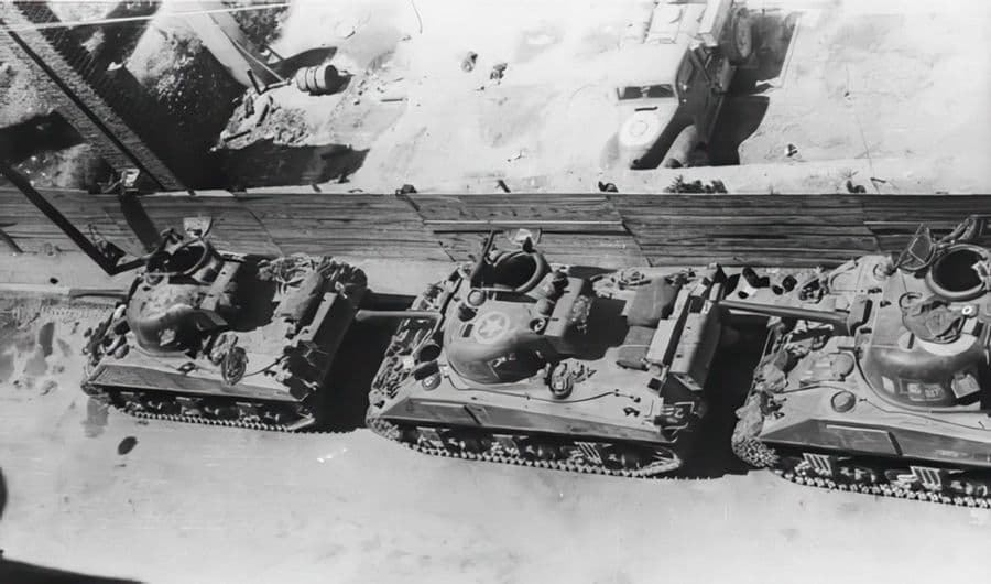 This image captures a moment in Trieste, Italy, in May 1945, featuring three Sherman III tanks belonging to the 20th Armoured Regiment of the 2nd New Zealand Division. The tanks are parked outside their billets on the Via Cicerone. From left to right